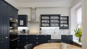 A modern kitchen with dark cabinetry, a wooden dining table, and a window with a view of the outdoors. - By The Sea (North Berwick)