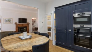 A dining area with a wooden table and chairs, a television above the fireplace, and a kitchen with dark blue cabinets and a built-in oven. - By The Sea (North Berwick)