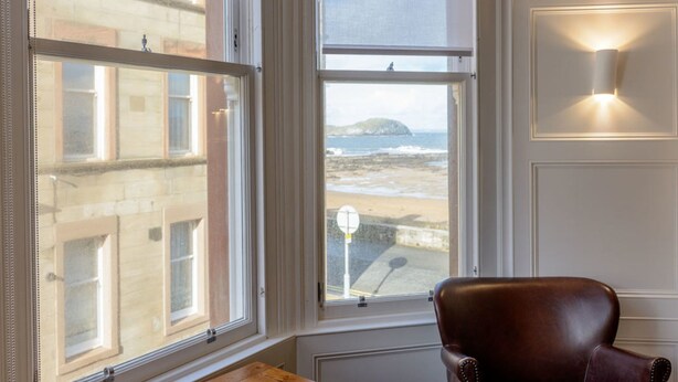 A room with a large bay window overlooking a beach, a leather armchair, and a wall-mounted light fixture. - By The Sea (North Berwick)