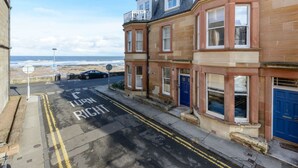 A coastal street with terraced houses, a clear sky, and a car parked on the side. - By The Sea (North Berwick)
