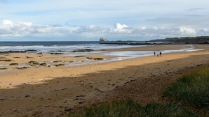 A beach with a rocky outcrop in the distance, sandy shore, and a few people walking. - By The Sea (North Berwick)