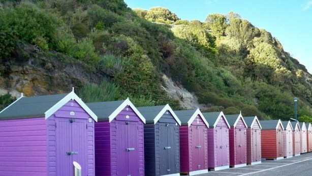 Una hilera de cabañas de playa con techos inclinados, alineadas en un estacionamiento con una colina al fondo.