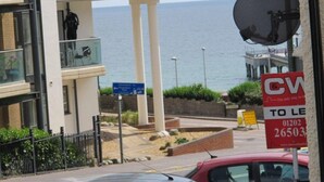 Un edificio moderno con balcón que da al mar, un cartel indicando que la propiedad está en venta y un coche rojo estacionado al frente.