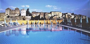A swimming pool with lounge chairs and umbrellas, surrounded by historic buildings and a stone wall. - Le Rocce (Sant'Agata de'Goti)
