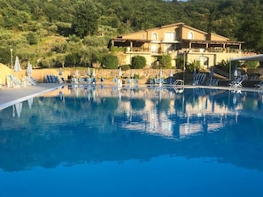 Une piscine entourée de chaises longues et de parasols, elle-même bordée par une colline et un bâtiment doté de nombreux balcons.