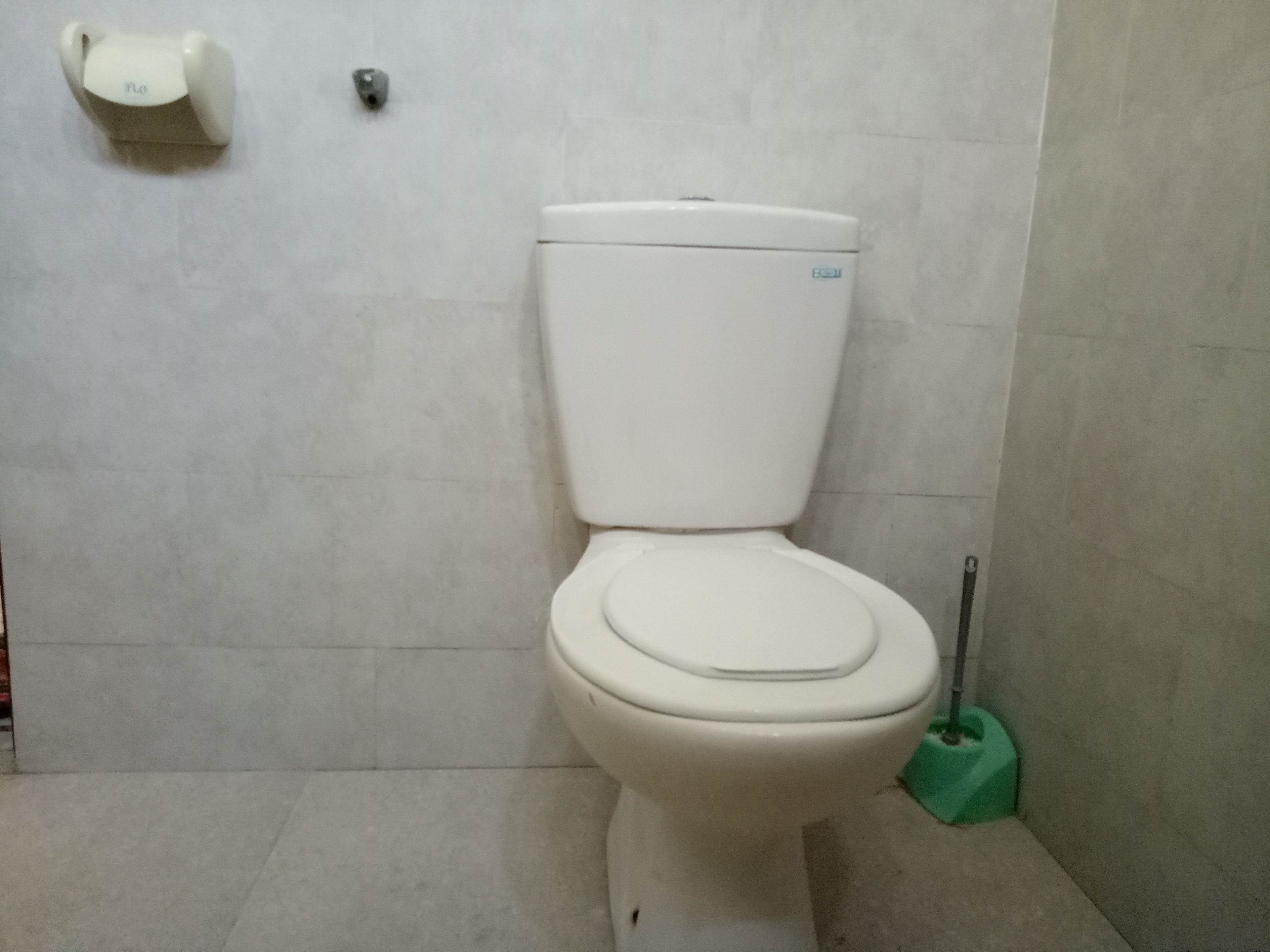 Un inodoro blanco con un cepillo de limpieza verde a un lado, en un baño con paredes de azulejos.