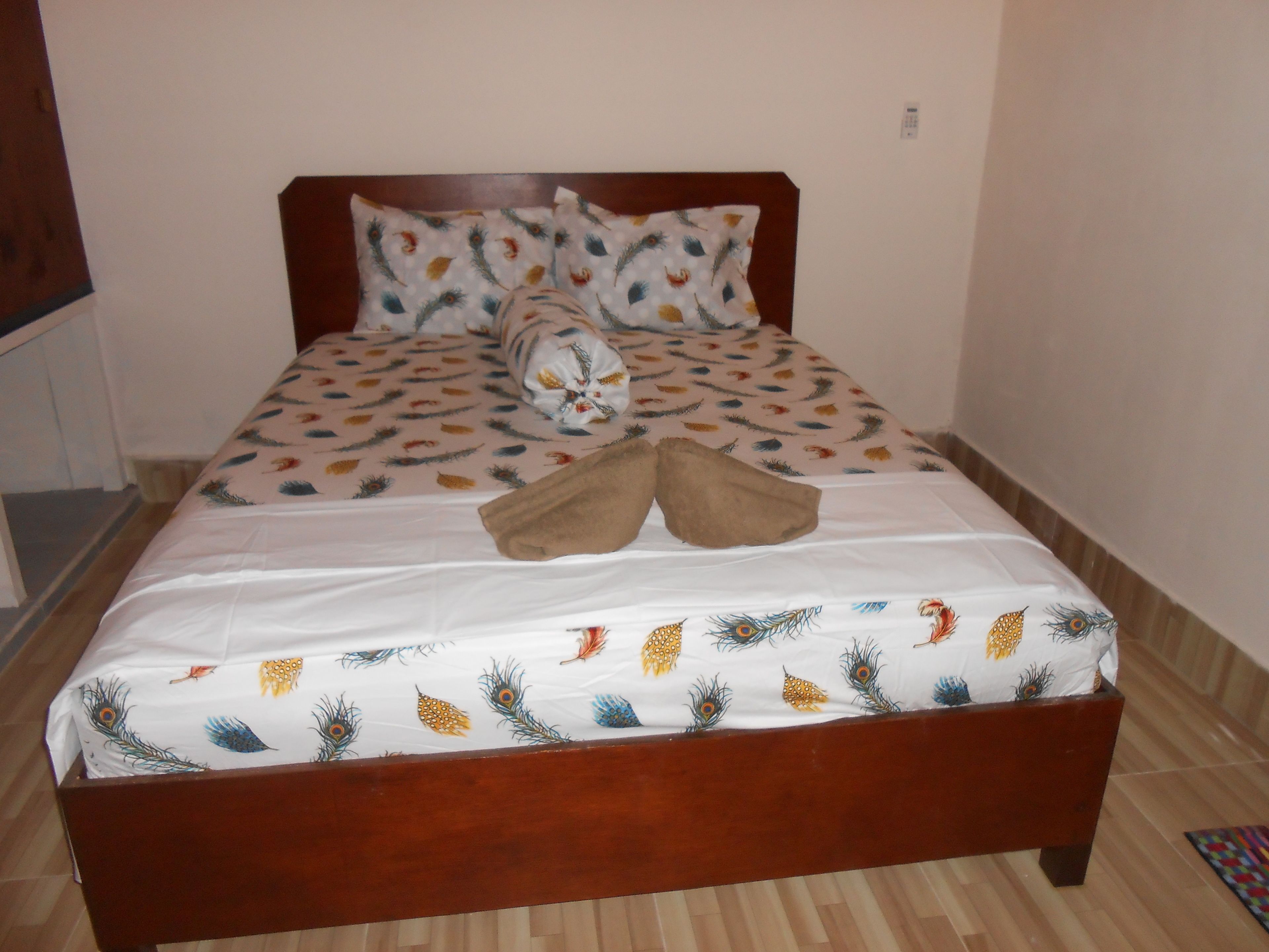 A bed with a white sheet featuring a peacock feather pattern, two pillows with a similar design, and a pair of brown slippers placed on the bed.
