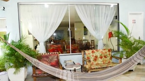 A room with a large window, a hammock, two chairs, a small table, and potted plants. - Pousada Âncora de Ouro (Cabo Frio)