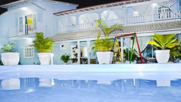 A pool area with a clear blue pool, white potted plants, and a building with balconies and a tiled roof. - Pousada Âncora de Ouro (Cabo Frio)