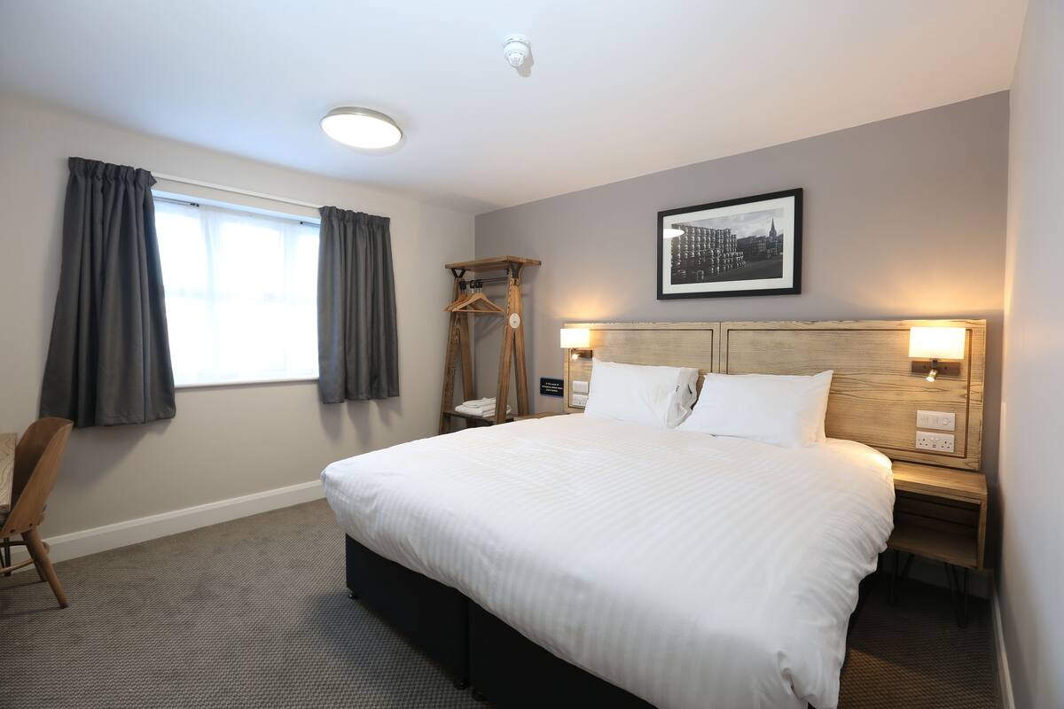 A hotel room with a large bed, two bedside tables, a wooden chair, a coat rack, and a framed picture on the wall.