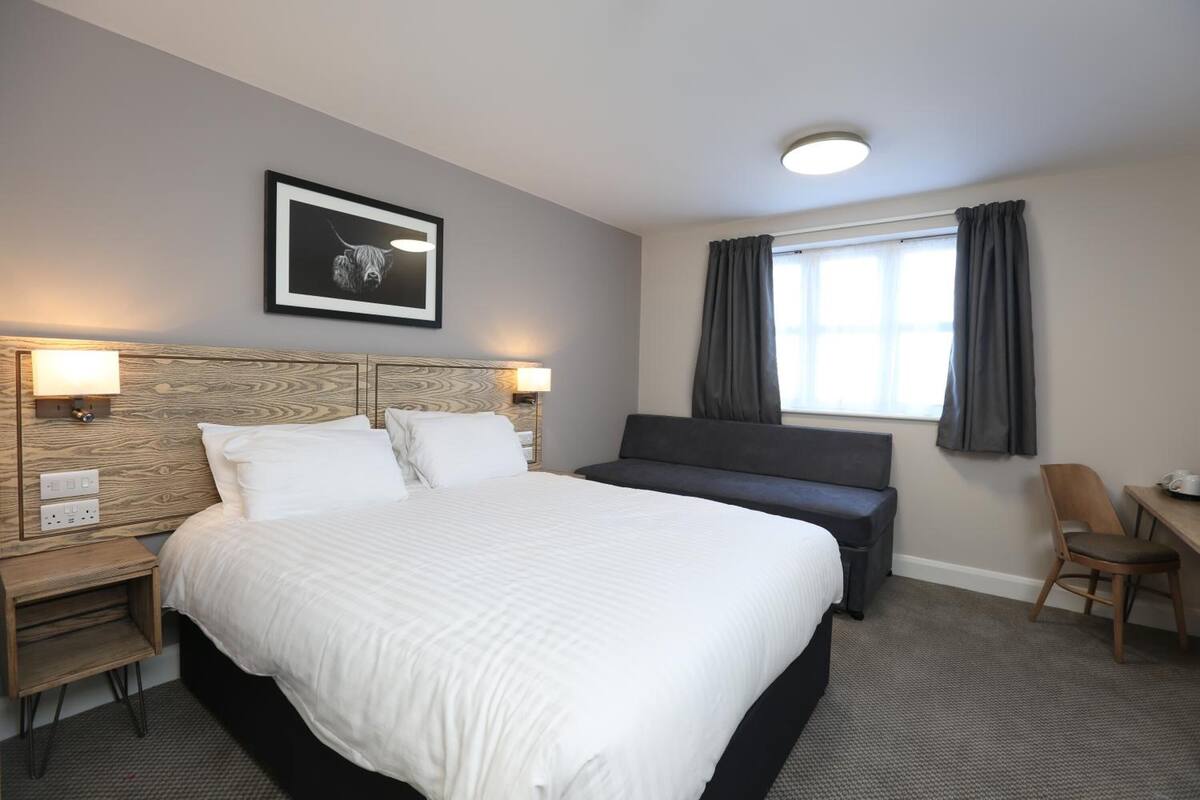 A hotel room with a large bed, a desk, a chair, a nightstand, a lamp, a framed picture on the wall, and a window with curtains.