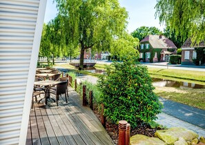 A wooden deck with outdoor furniture, a pond, and residential buildings in the background. - Hotel-Restaurant Hilling (Papenburg)