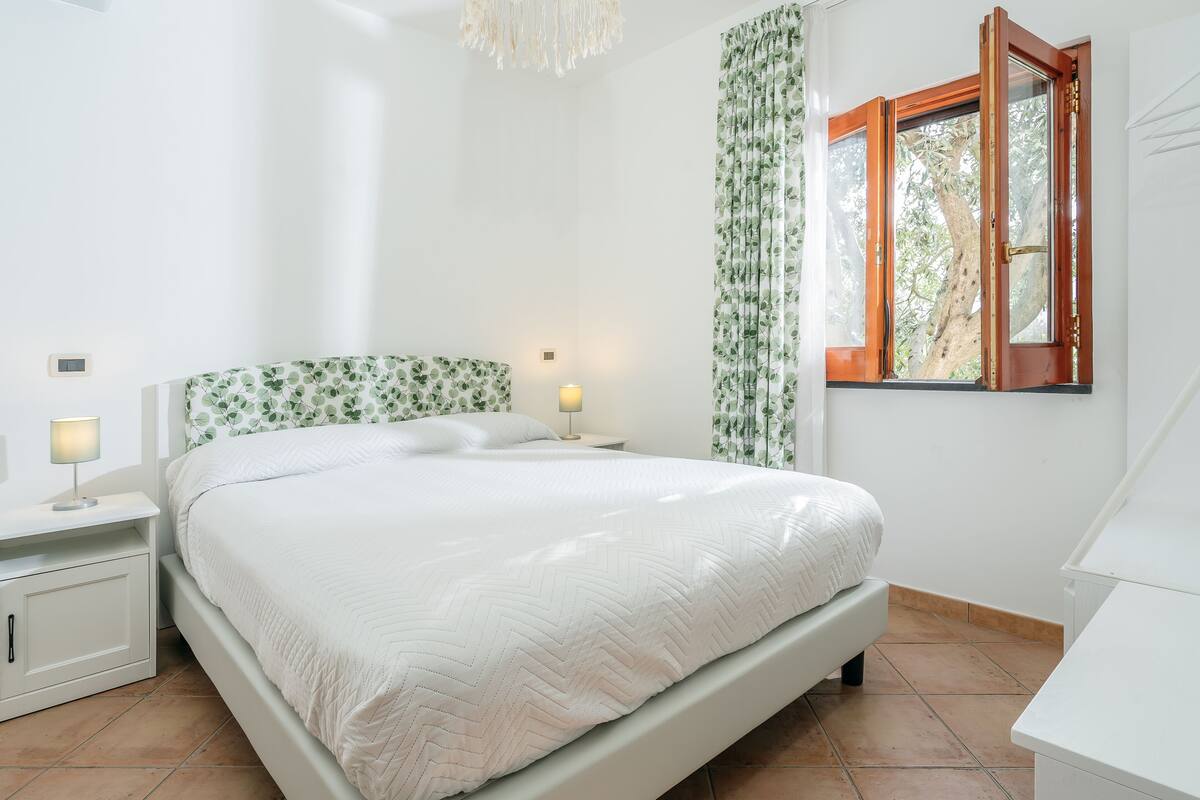 A bedroom with a bed, nightstand, lamp, and window with curtains.