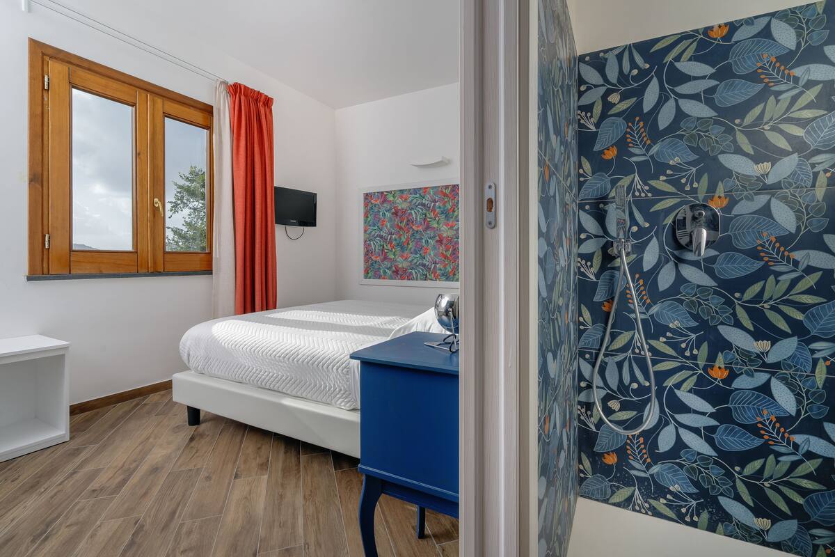 A bedroom with a bed, a blue dresser, a shower with a floral tile wall, and a window with curtains.