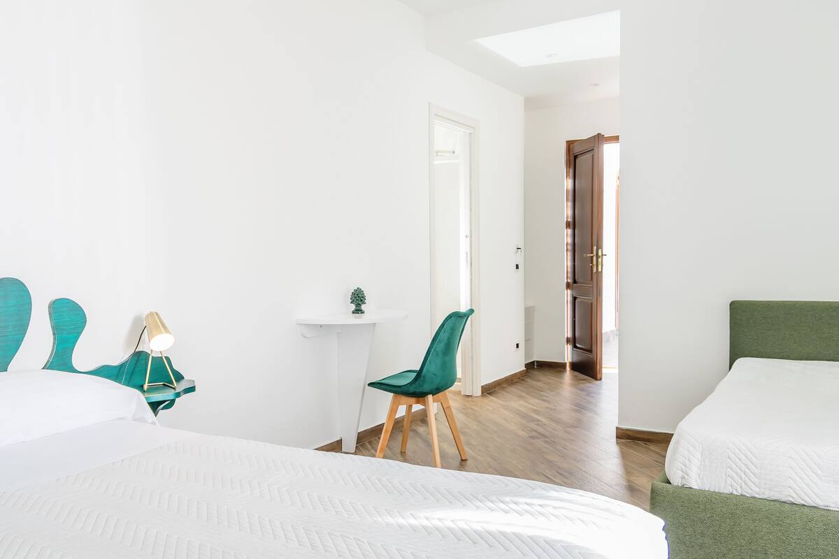 A hotel room with two beds, a desk, a chair, and a small table with a lamp.