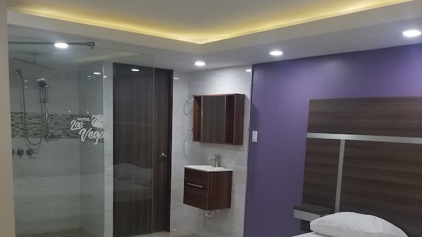 A hotel room with a bed, a shower area, a vanity with a mirror, and a purple accent wall.