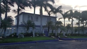 A two-story building with a white exterior and a blue roof, surrounded by palm trees and a white fence.