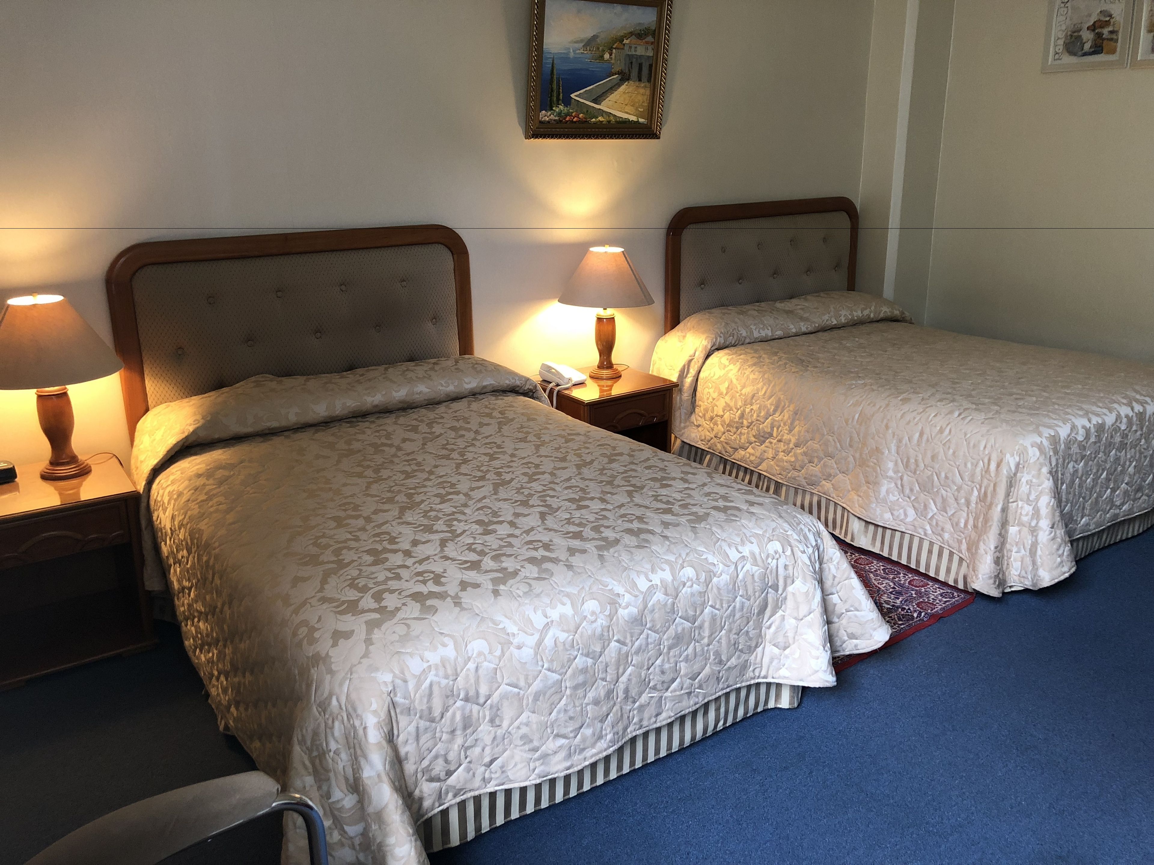 A hotel room with two beds, each with a headboard, bedside lamps, and a nightstand. A painting is on the wall above the beds.