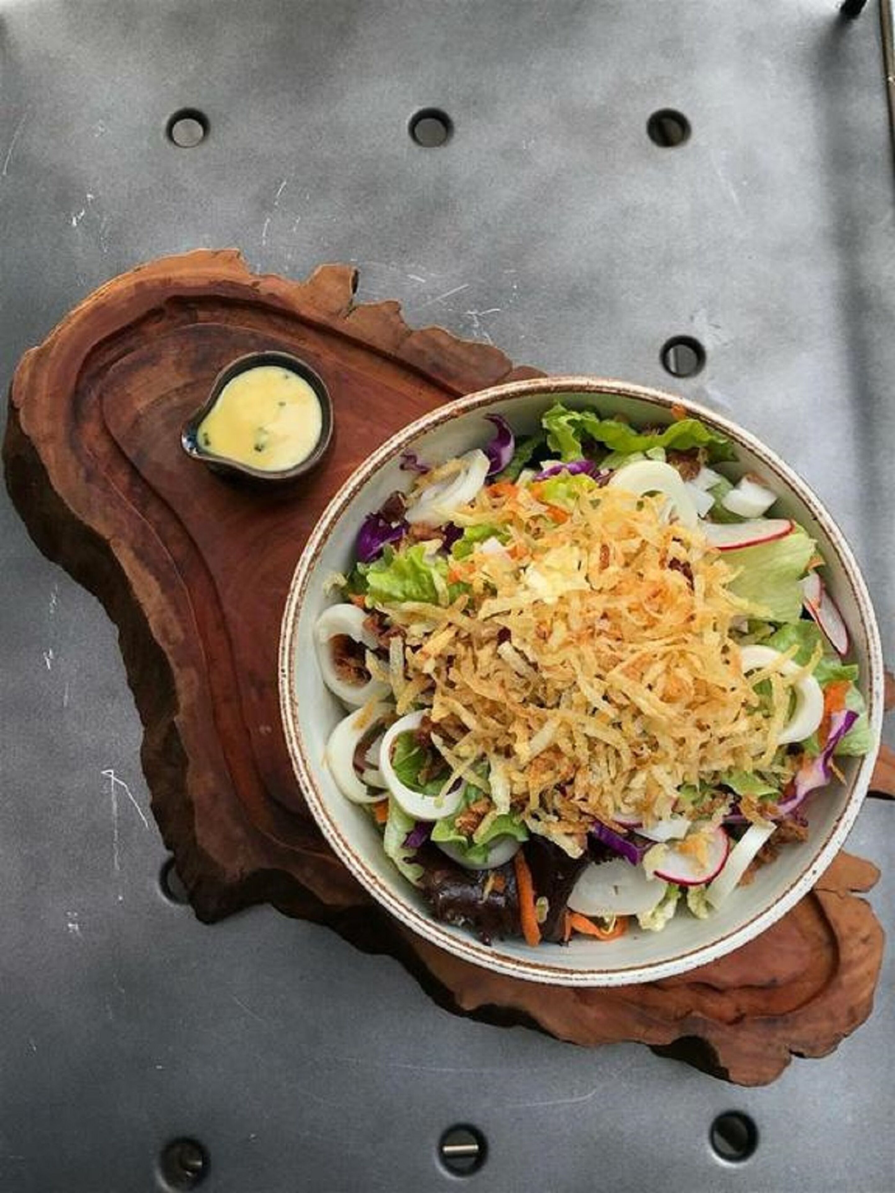 A bowl of salad with shredded toppings on a wooden board, accompanied by a small bowl of dressing.