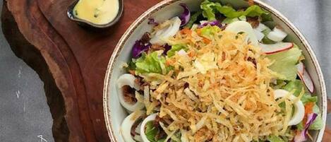A bowl of salad with shredded toppings on a wooden board, accompanied by a small bowl of dressing.