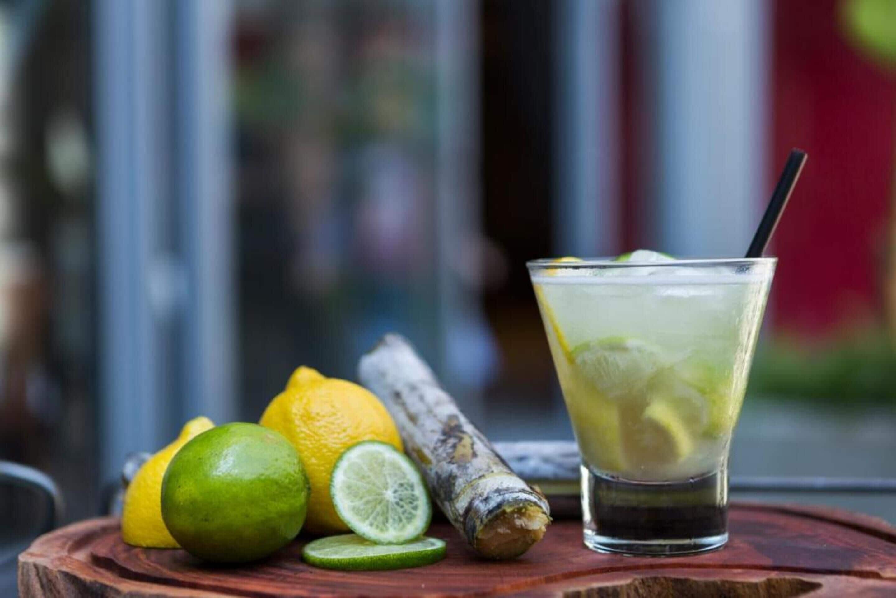 Un vaso de bebida helada con rodajas de lima y una pajita, acompañado de limas enteras y cortadas, y un trozo de jengibre sobre una superficie de madera.