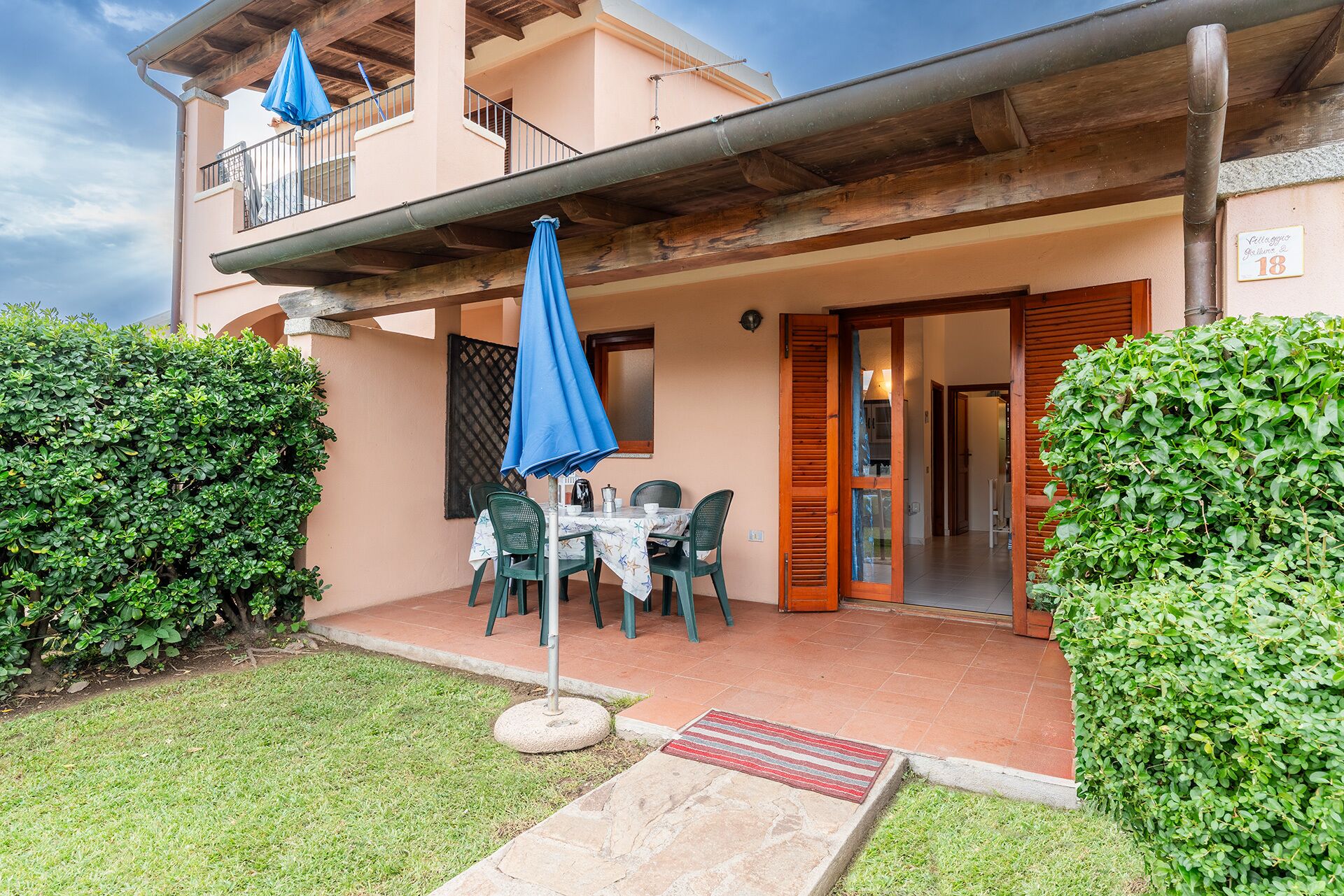 A patio with a table and chairs, a blue umbrella, and a wooden door leading inside.