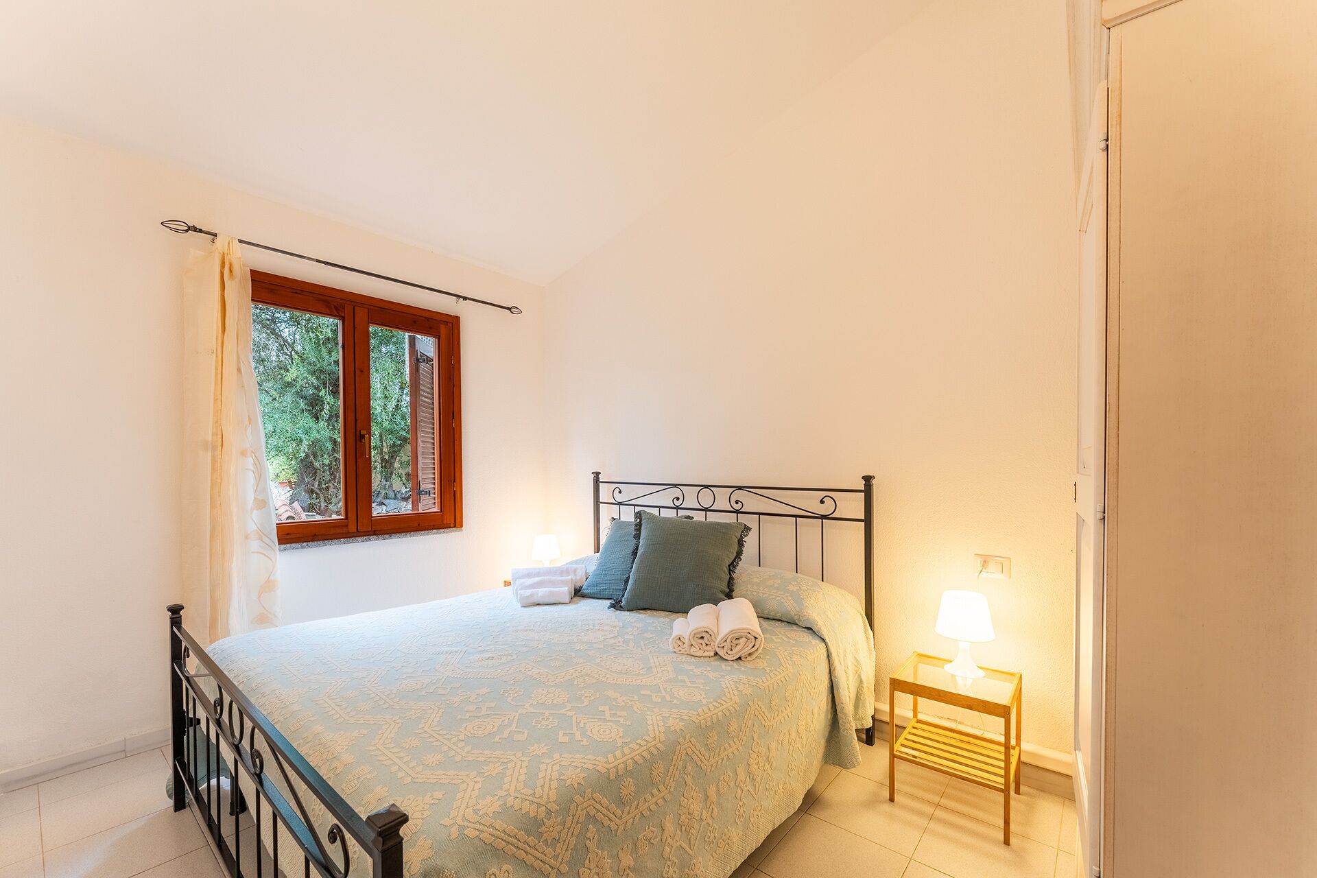 A bedroom with a bed, a window, a bedside table with a lamp, and a door.