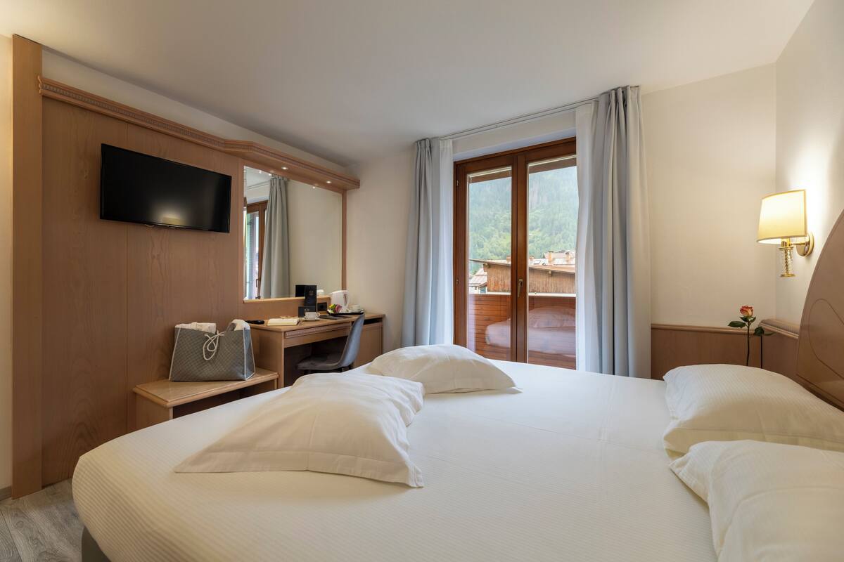 A hotel room with a large bed, a desk with a computer, a TV mounted on the wall, and a window with curtains.