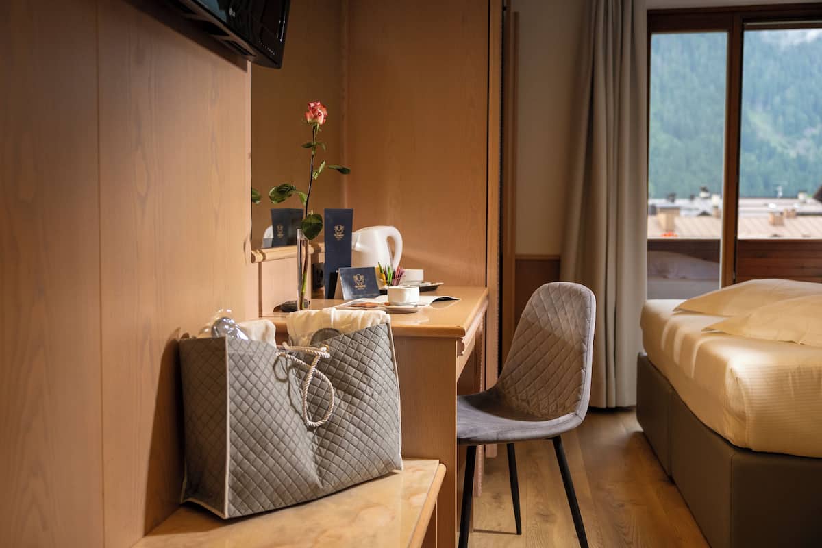 A hotel room with a wooden desk, a chair, a bed, a window with a mountain view, and a vase with a flower.