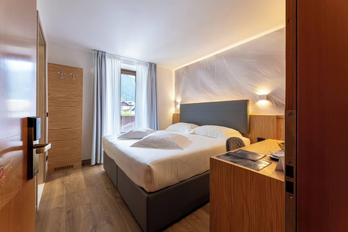 A modern hotel room with a large bed, a wooden wardrobe, a desk with a laptop, and a view of the outdoors.
