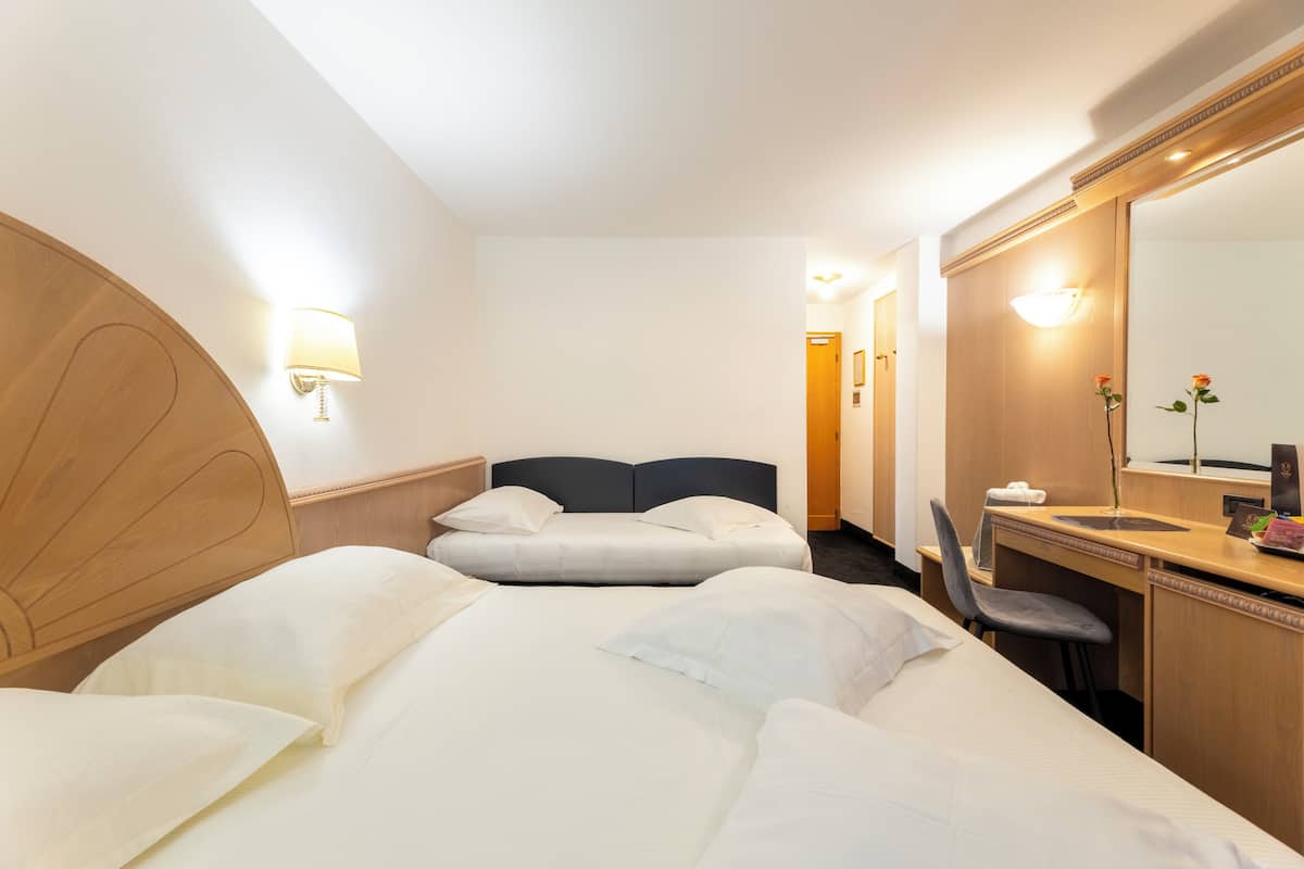 A hotel room with a large bed, a desk with a chair, a mirror, and a wall-mounted lamp.