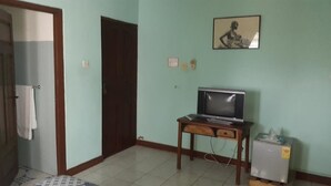 Una habitación con piso de baldosas, un escritorio de madera con televisor, un refrigerador pequeño y una puerta que da a otra habitación.