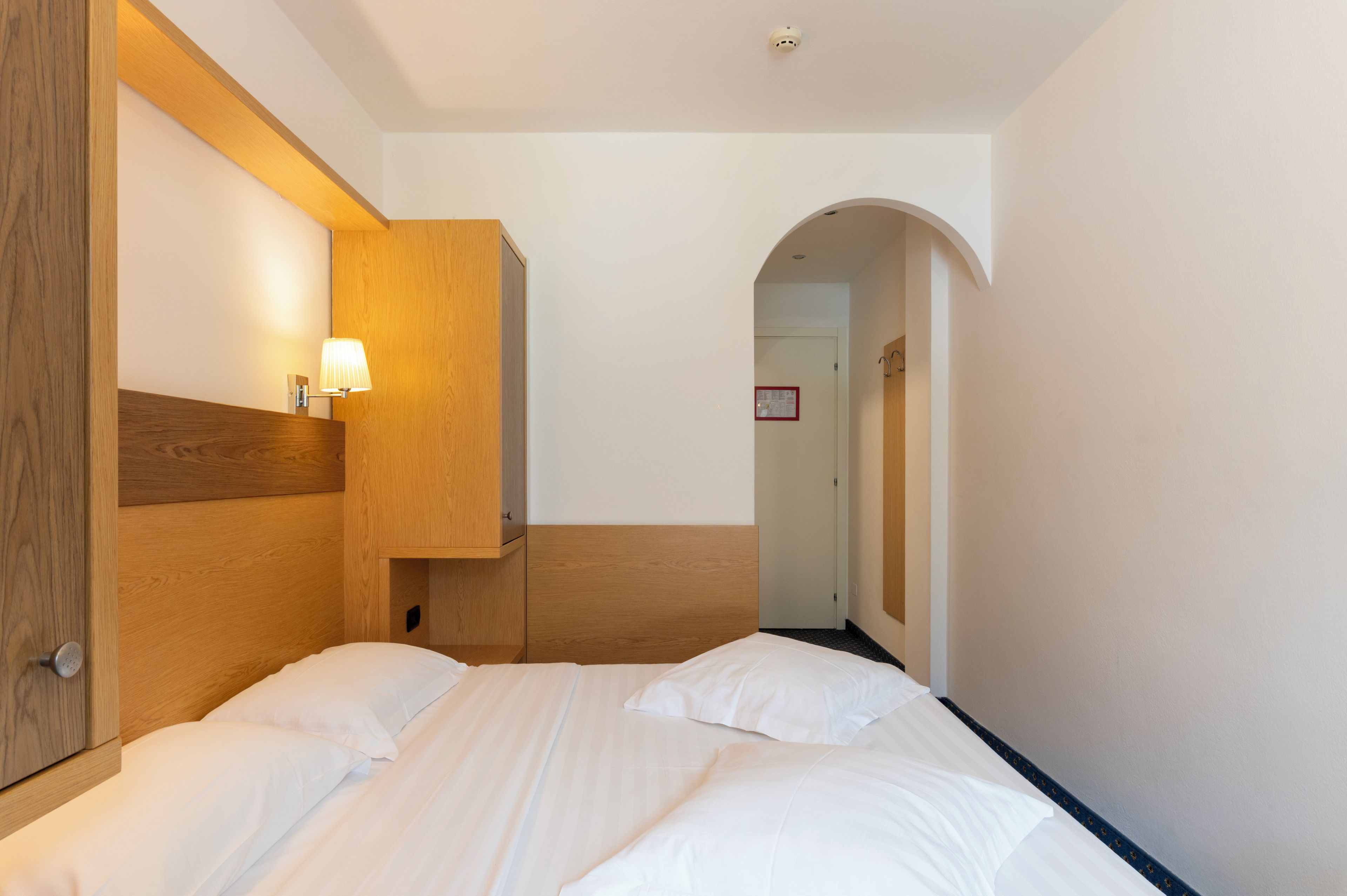 A hotel room with a bed, a wooden headboard, a wall-mounted lamp, and a door with a sign.