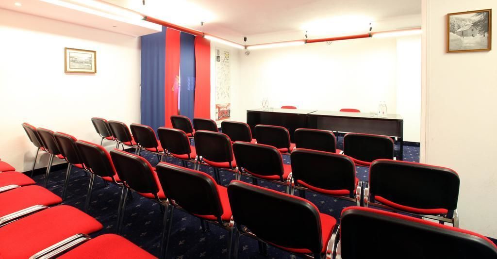 A conference room with rows of red chairs, a table with a microphone, and a blue curtain.