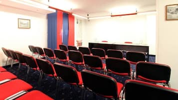 A conference room with rows of red chairs, a table with a microphone, and a blue curtain.