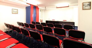 A conference room with rows of red chairs, a table with a microphone, and a blue curtain.
