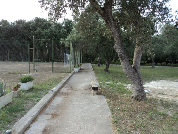 Un sendero de concreto bordeado por árboles y una zona cercada con un arco de fútbol.