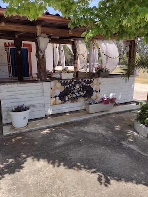 Un quiosco rústico al aire libre con un letrero que dice 'Happy Birthday', estructura de madera encalada y plantas en macetas.