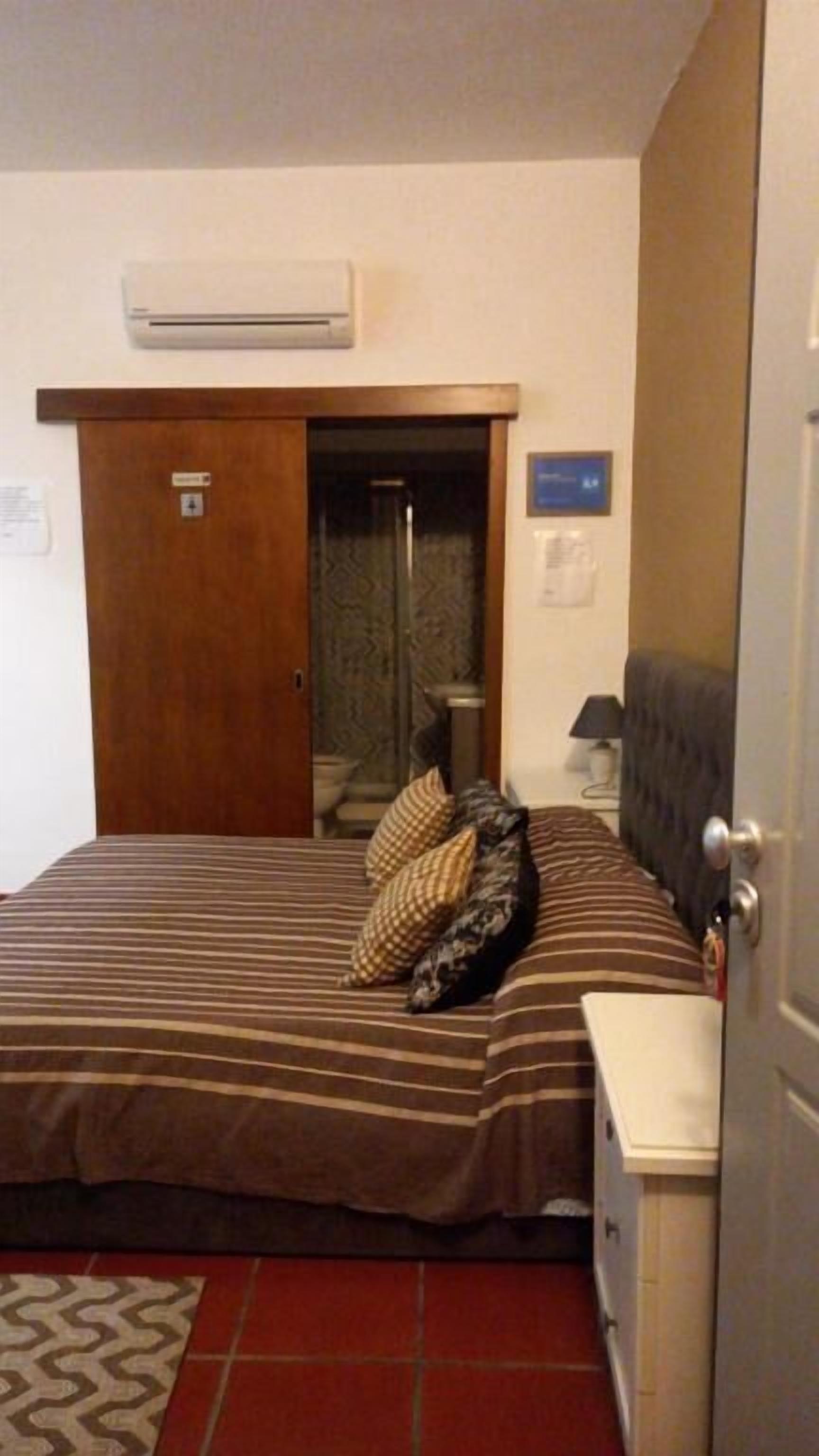 A hotel room with a bed, a bedside table, an air conditioning unit, a door, and a bathroom visible through an open door.