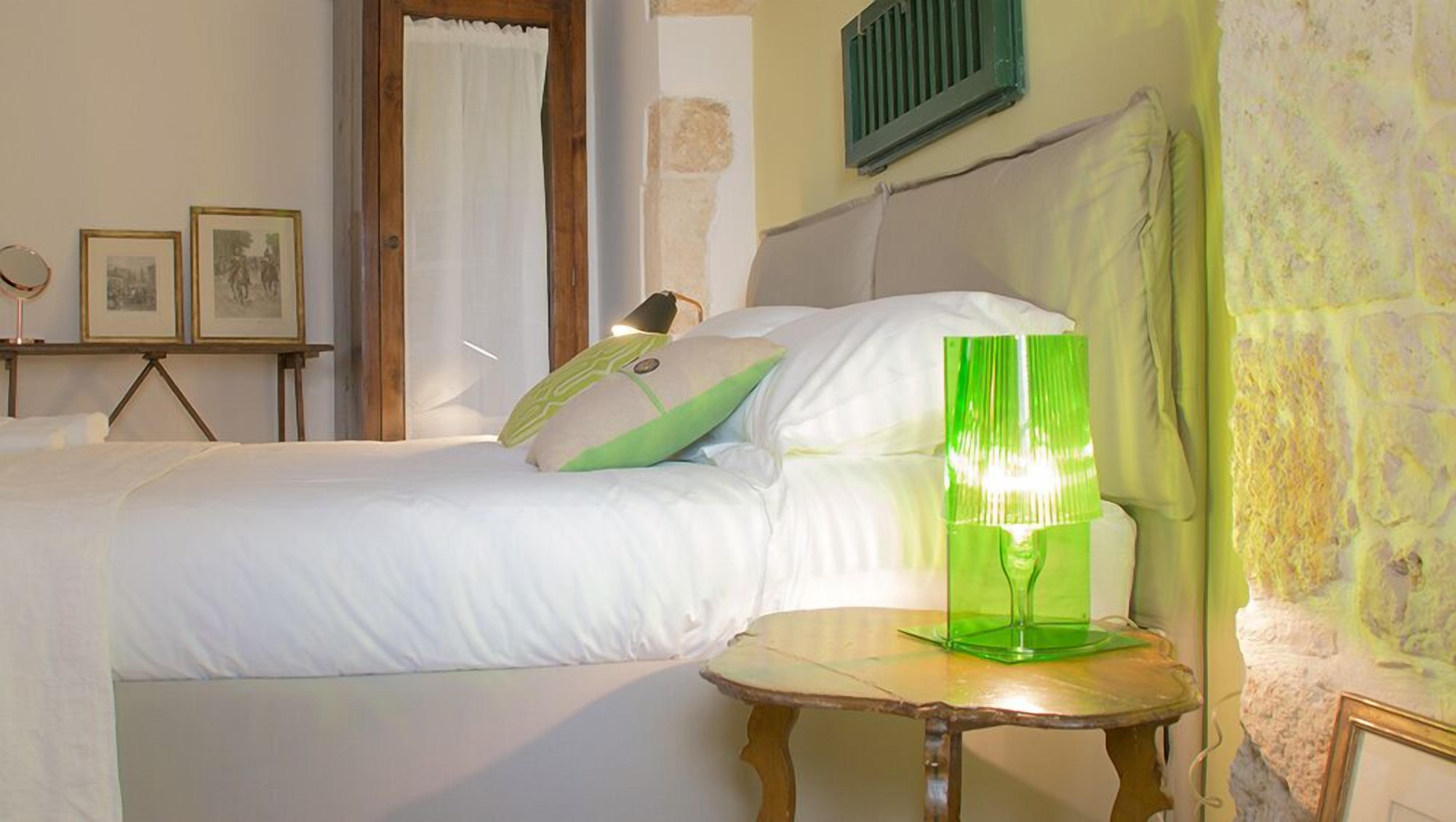Un dormitorio con cama, almohadas, una mesita de noche con una lámpara verde y cuadros en la pared.