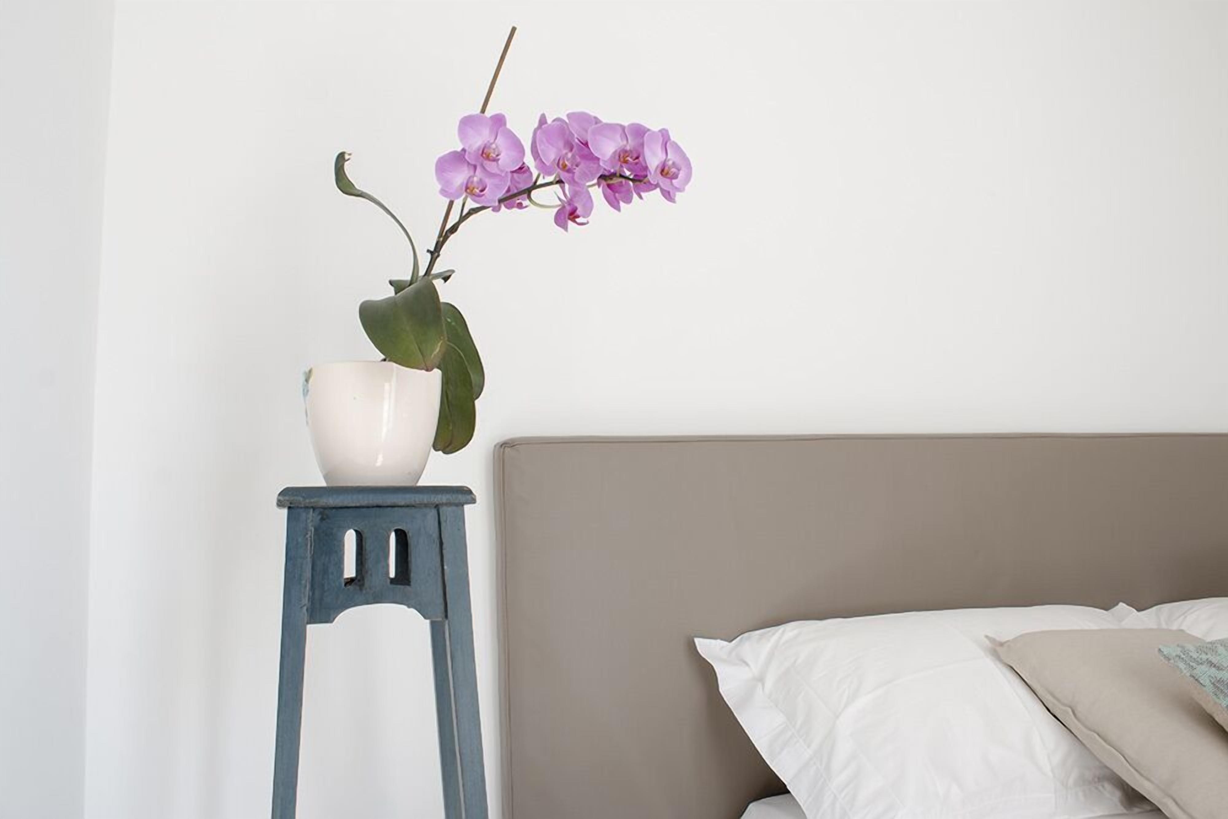 A bed with a beige headboard, a white pillow, and a small blue stool with a white vase containing purple flowers.