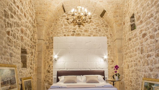 Habitación de paredes de piedra con cama, candelabro, cuadros enmarcados y una pequeña mesa con planta en maceta.