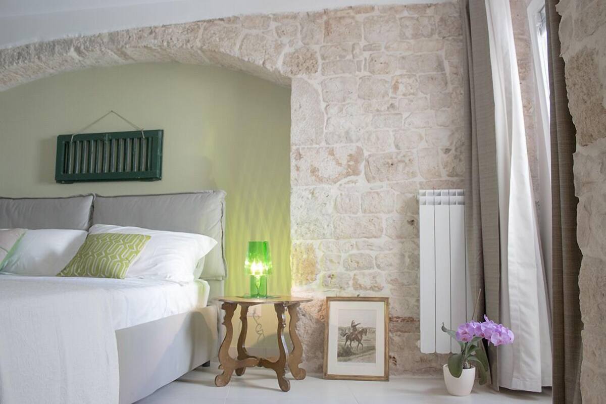 A bedroom with a stone wall, a bed with white bedding, a green lamp, a framed picture, and a potted plant.