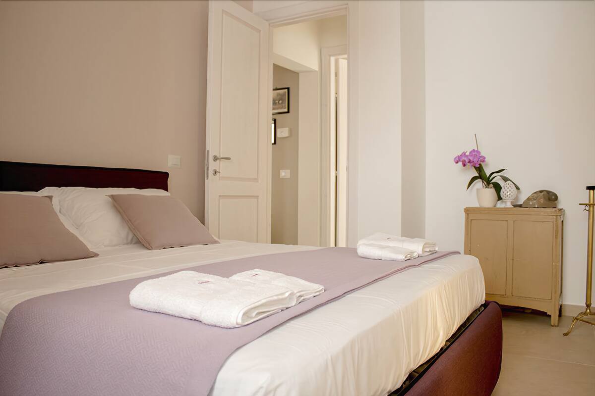 A neatly made bed with white and beige bedding, a bedside table with a vase of flowers, and a door leading to another room.