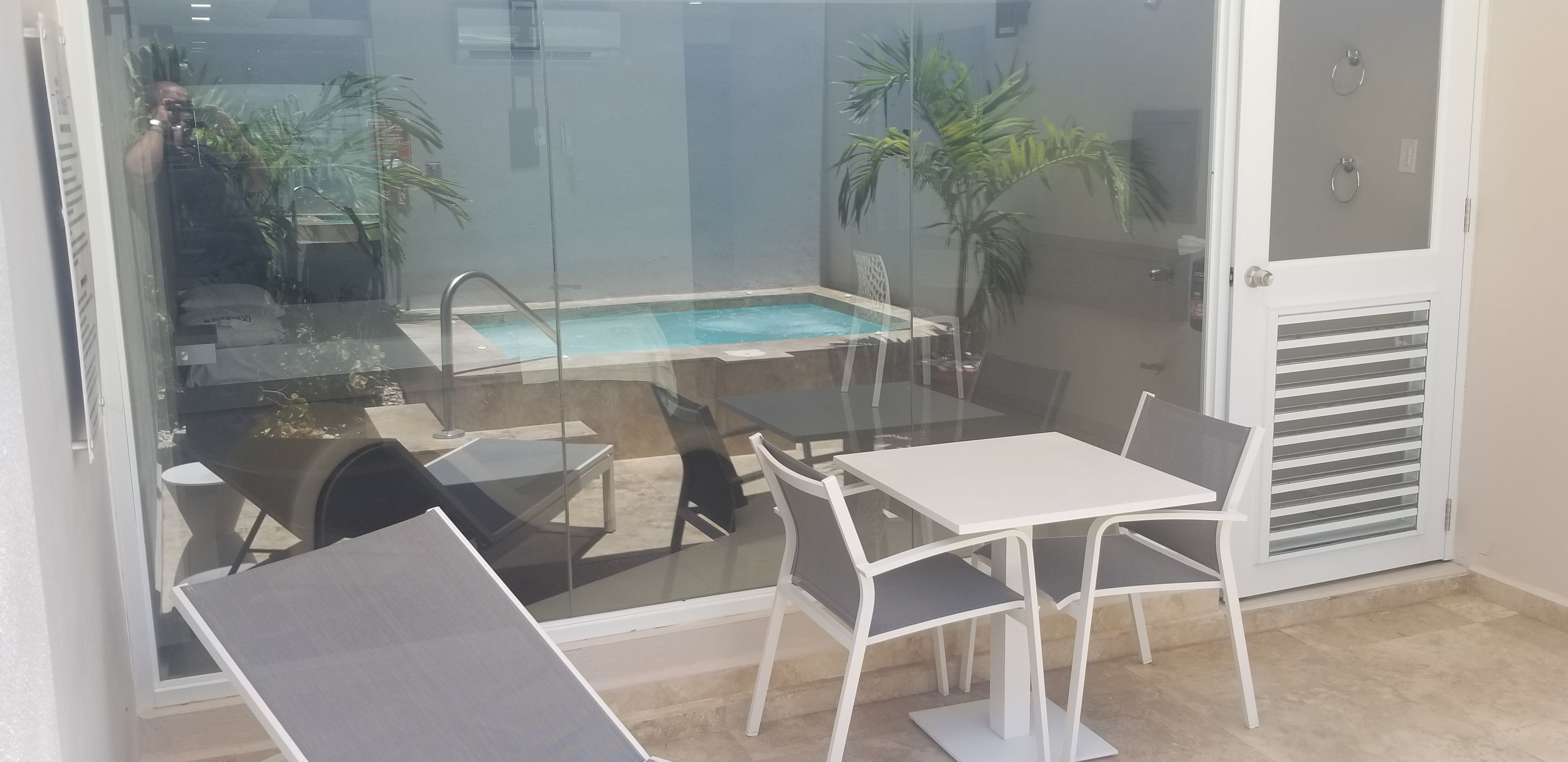 A glass-enclosed balcony with a pool, white tables and chairs, and a door with a vent.