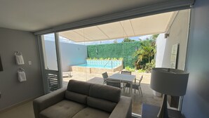 Sala de estar moderna com sofá, mesa e luminária. Há uma porta de vidro deslizante que dá para uma varanda com piscina e vegetação.