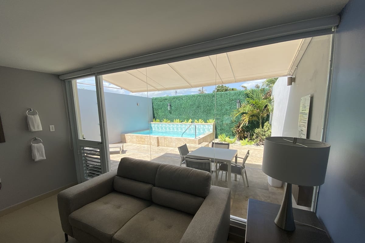 A modern living room with a sofa, a table, and a lamp. There is a sliding glass door leading to a balcony with a pool and greenery.