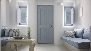 A room with a light blue door, two windows with curtains, a white table with a candle, and a blue sofa with striped pillows. - Palmariva Villas (Santorini)
