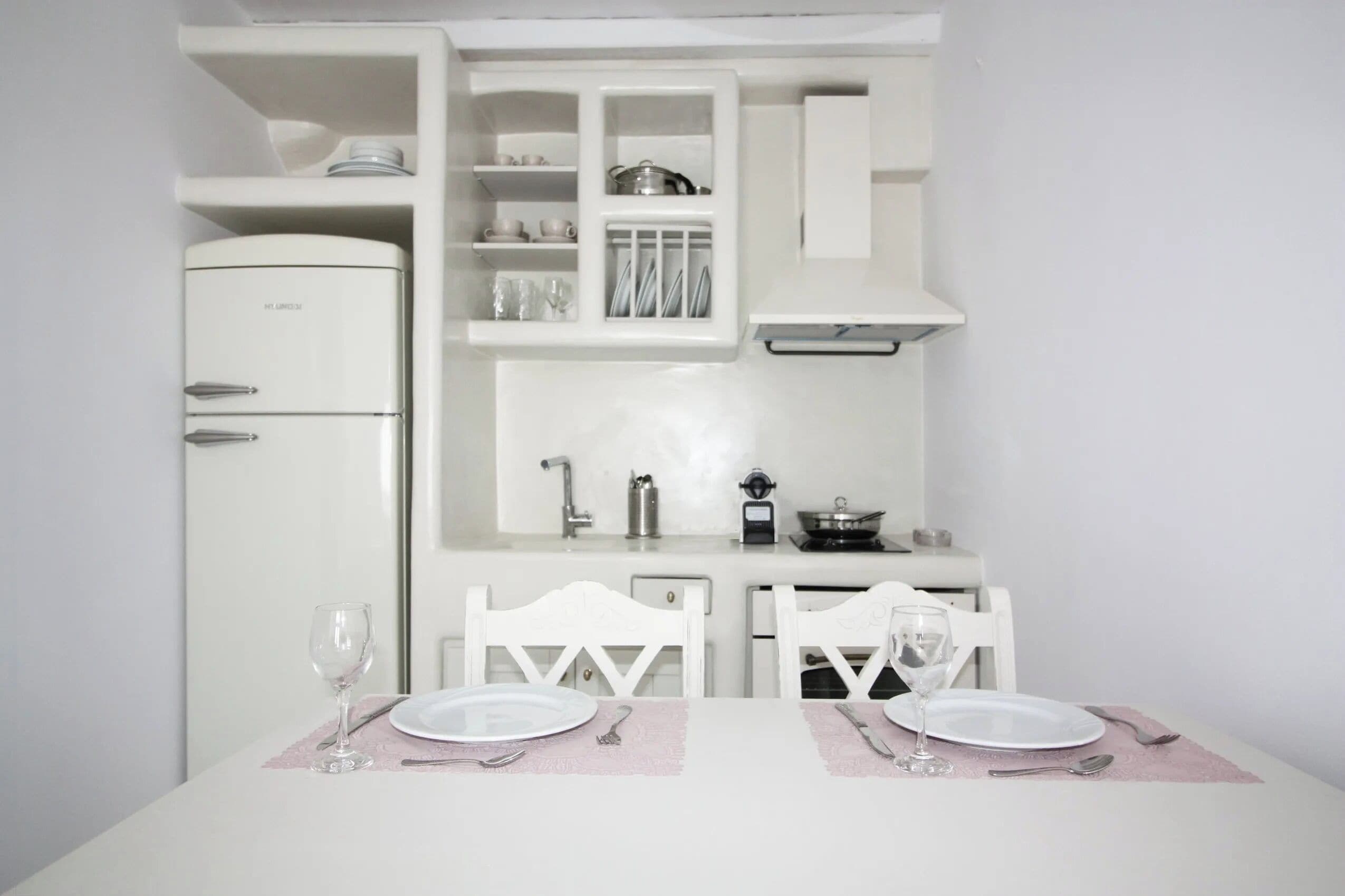 Cocina compacta con armarios blancos, horno empotrado y mesa de comedor para dos personas con sillas blancas.