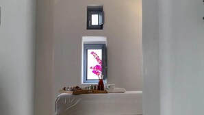 A narrow hallway leading to a bedroom with a bed, a window with a view, and a small shelf with items. - Palmariva Villas (Santorini)